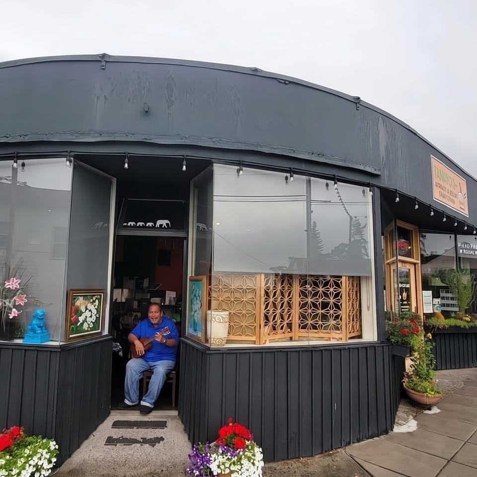 A man playing a ukulele sits in the open doorway of a dark gray restaurant with large windows.