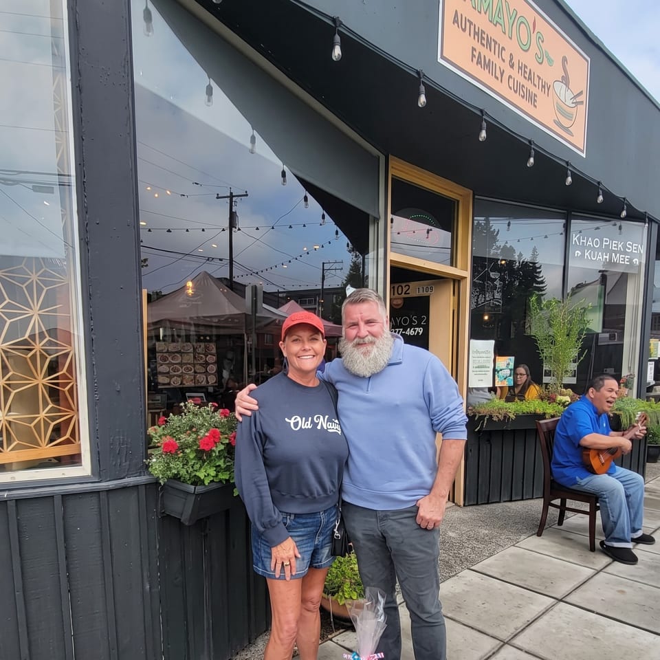A couple stands outside Mayo's restaurant, with a man playing a ukulele nearby.