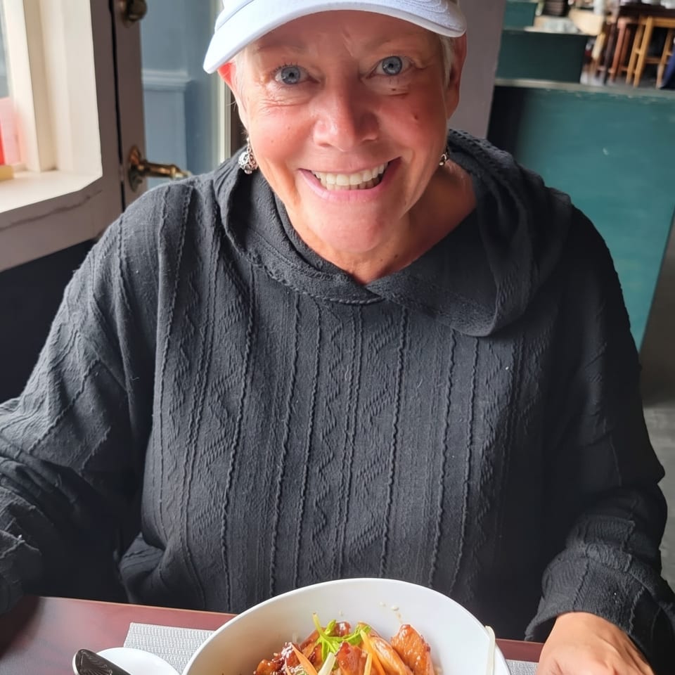 A smiling woman in a white cap and black hoodie sits at a table with a bowl of food.